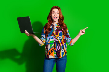 Wall Mural - Young woman with red hair holding a laptop on green background while smiling and pointing sideways