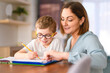 © Louis-Paul Photo - homework and school concept - smiling student boy with book writing to notebook at home with his mother help her