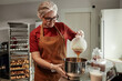 © Seventyfour - Medium shot of young female baker wearing hairnet pouring maple syrup into metal bowl mixing ingredients while baking artisan bread in bakehouse kitchen