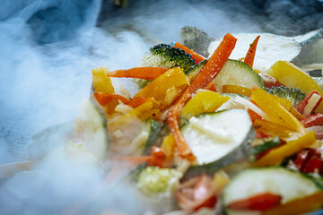 A steaming vegetable stir-fry with zucchini, bell peppers, carrots, and broccoli is being cooked in a pan, releasing aromatic steam. The vibrant colors highlight the freshness of the ingredients.