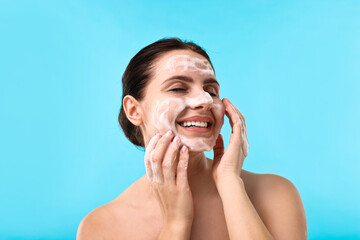 Wall Mural - Smiling woman washing her face with cleansing foam on light blue background