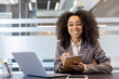 © Tetiana - Portrait of a smiling young African-American businesswoman sitting at a desk in the office with a laptop, holding a notebook and a pen in her hands, looking confidently at the camera