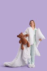 Wall Mural - Young woman in pajamas with pillow, blanket and teddy bear on lilac background