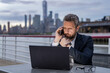 © Volodymyr - Businessman working on laptop talking on phone outdoor. Senior boss working on laptop outside. Mature businessman with remote work. Handsome business man using laptop computer.