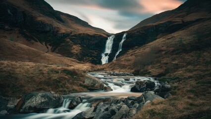 Wall Mural - Cascading waterfall flows through rocky valley surrounded by dark hills under a moody sky with soft pastel colors in the background.