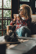 © simona - Mature woman sitting alone at home holding a cup of hot herbal tea while looking around with a relaxed expression enjoying a moment of self-care balance and peace in a cozy environment filled