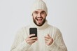 © mutter - The image depicts a cheerful young bearded man wearing a grey T-shirt and a fur cap hat, overcome with surprise and joy, as he showcases a winning gesture on his smartphone set against a plain white