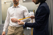 © ADDICTIVE STOCK - Tailor measuring a man sleeve for a custom shirt fitting in suit store