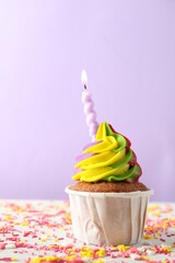 Wall Mural - One delicious cupcake with colorful cream, burning candle and sprinkles on white table against violet background, closeup