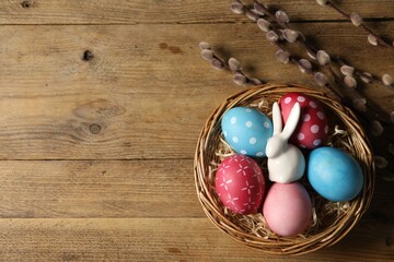 Wall Mural - Beautiful decorated Easter eggs in wicker bowl, bunny figure and willow branches on wooden table, flat lay. Space for text