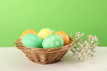 Wall Mural - Many decorated Easter eggs in wicker basket and gypsophila flowers on wooden table against light green background, closeup