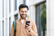 © Liubomir - Young student with backpack on shoulder using app on smartphone. Man walking outside university campus smiling with phone in hands.