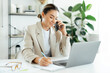 © Kateryna - Smiling pretty caucasian business woman, company manager talking on the mobile phone and writing notes at her desk with a laptop in a bright modern office, having smartphone conversation with client