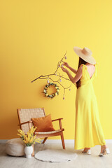 Wall Mural - Young woman hanging Easter eggs on tree branches near yellow wall in room, back view