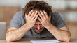 © JKLoma - Overwhelmed: A man is hunched over a desk with a look of distress on his face, a symbol of stress and struggle
