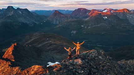 Wall Mural - couple hiking in norway man and woman friends raised hands enjoying aerial sunset mountains view, ac