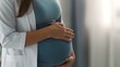 © Tanakrit - A pregnant woman gently touches her belly while wearing a teal top and a white coat, conveying a sense of care and anticipation.