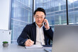 © RomArt production - Confident businessman in formal attire discussing over phone call while taking notes in a notepad. Situated in a bright, glass-walled modern office space enhanced with natural light.