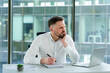 © RomArt production - Professional man sitting in a modern office, wearing formal attire, writing on a notebook, displaying focused and pained expressions. Bright workspace with windows and a laptop on a white desk.