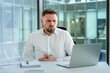 © RomArt production - Male professional in modern office dressed casually facing discomfort while using a laptop, expressing stress due to an internal issue. The setting reflects a corporate environment with large windows.