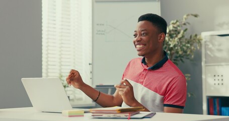 Wall Mural - Dark-skinned boy in t-shirt is finished talking on phone satisfied with service products sold monthly profits sits in front of computer victory dance, hands clenched into fists looks at camera, smiles