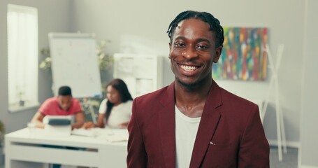 Wall Mural - Portrait of handsome elegant man with dreadlocks and stubble dark skin, looks into camera with brown eyes, smiles broadly showing straight white teeth, wearing a maroon jacket and white tshirt