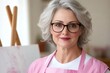 © Sanchai - Attractive cheerful female pensioner in apron and eyeglasses imroving artistic skills, standing in studio next to easel, holding paintbrush during art workshop People, leisure and hobby concept