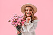 © HBS - Portrait of a beautiful young woman in a beige spring dress and hat holding a bouquet of pink flowers and laughing. Concept of spring, fashion, cheerful mood. International Women's Day. 8 March.