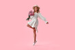 © HBS - Full length image of a young stylish smiling cheerful beautiful woman in a beige dress and hat holding a bouquet of pink flowers and jumping on a soft pink background. Advertising concept.