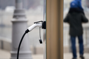 A person is walking by a charging station for electric cars. The person is wearing a black coat and a blue hat