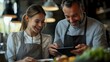 © Yuliia - Cheerful Caucasian female and male chefs in aprons looking at digital tablet in restaurant kitchen, sharing professional culinary experience.