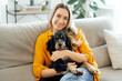 © Kateryna - Cheerful caucasian charming woman in a casual wear, sitting on a soft couch in a living room, embracing her lovely dog, looking at the camera with a happy expression, enjoying a cozy moment at home