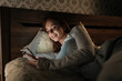 © dikushin - Cheerful young woman relaxing in bed, scrolling smartphone with soft bedroom lighting, enjoying late night digital connection and personal downtime. Concept of mobile addiction and insomnia.