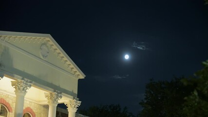 Wall Mural - The moon shines over the white columned house in the evening with a full moon. Dark sky above has a visible moon with soft glow. Tree branches frame the top of the image. The atmosphere feels calm and