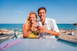 © Jose Calsina - Joyful couple toasting with drinks on a sunny beach, enjoying refreshing beverages while capturing the perfect moment by the shoreline under a clear blue sky filled with warm sunshine and laughter