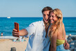 © Jose Calsina - A Joyful Young Couple Taking Selfie at the Beach with Smartphone on a Beautiful Sunny Day. Boyfriend and Girlfriend Create Lasting Memories Against the Stunning Ocean Waves at Coastline on Vacations