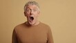 © olga_demina - Senior man with grey hair and brown sweater expressing shock and surprise, opening his mouth in amazement while standing against a beige background, conveying astonishment and disbelief