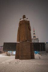 The Latvian Riflemen statue stands on a snowy pedestal in Riga, Latvia, with St. Peter's Church spire illuminated against a cloudy winter evening sky.