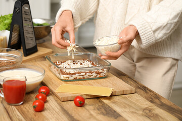 Wall Mural - Beautiful mature woman adding cheese to delicious lasagna at table in kitchen, closeup