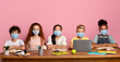 © Prostock-studio - Education during virus outbreak concept. Multinational schoolkids in medical masks sitting at desk over pink background, free space. Panorama