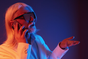 Wall Mural - Stylish elderly man talking on the phone with a modern attitude, wearing sunglasses, dressed in a white suit, with a colorful gradient background featuring blue and pink tones