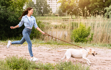 active pet concept. laughing girl chasing their running dog in park near lake, copy space, full leng