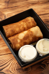 Wall Mural - Delicious crepes served with creamy dipping sauces in a takeout box on a wooden table