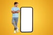 © Prostock-studio - Cheerful smiling casual guy standing with crossed hands looking at giant big cell phone, empty white screen template, orange studio wall. Mock up for app or website, free copy space, full body length