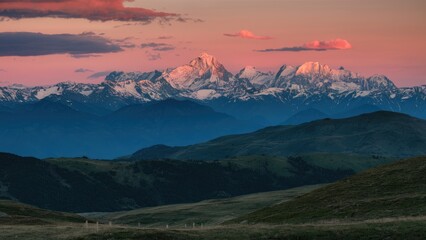 Wall Mural - Majestic mountain sunset with vibrant pink and orange clouds over snow-capped peaks, deep blue and green hills in the foreground, serene landscape.