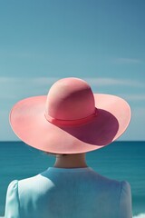 Wall Mural - a woman wearing a wide-brimmed pink hat, viewed from the back, with a sky-blue sea in the background