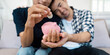 © itchaznong - Diverse LGBTQIA couple saving money together, placing coins into a pink piggy bank at home.