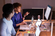 © Studio Marmellata - Two young men sit at a desk in a dimly lit office, discussing financial data displayed on multiple screens while analyzing stock market trends and investment opportunities.