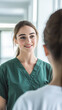 © Studio 70SN - Healthcare nurse junior doctor hospital corridor young woman Caucasian brunette hair long, green scrubs talking with patient smiling, window light