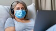 © A4719 - A person wearing a face mask and headphones, using a laptop while reclining on a comfortable pillow. Gray shirt, soft lighting, indoor setting